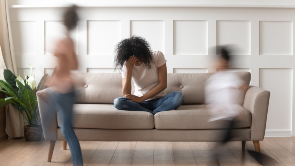 Crianças correndo enquanto a mãe está sentada no sofá, apresentando bastante irritabilidade, um dos sintomas de esgotamento mental.