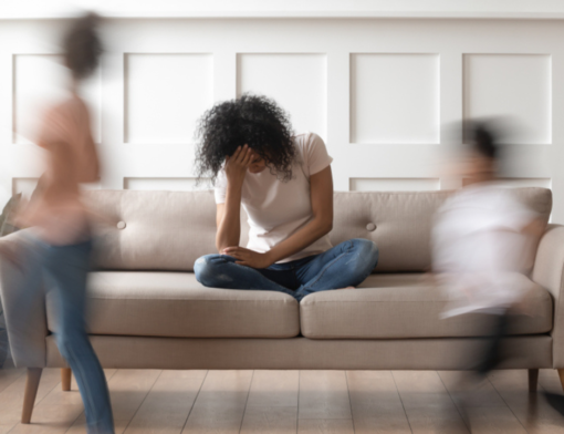 Crianças correndo enquanto a mãe está sentada no sofá, apresentando bastante irritabilidade, um dos sintomas de esgotamento mental.