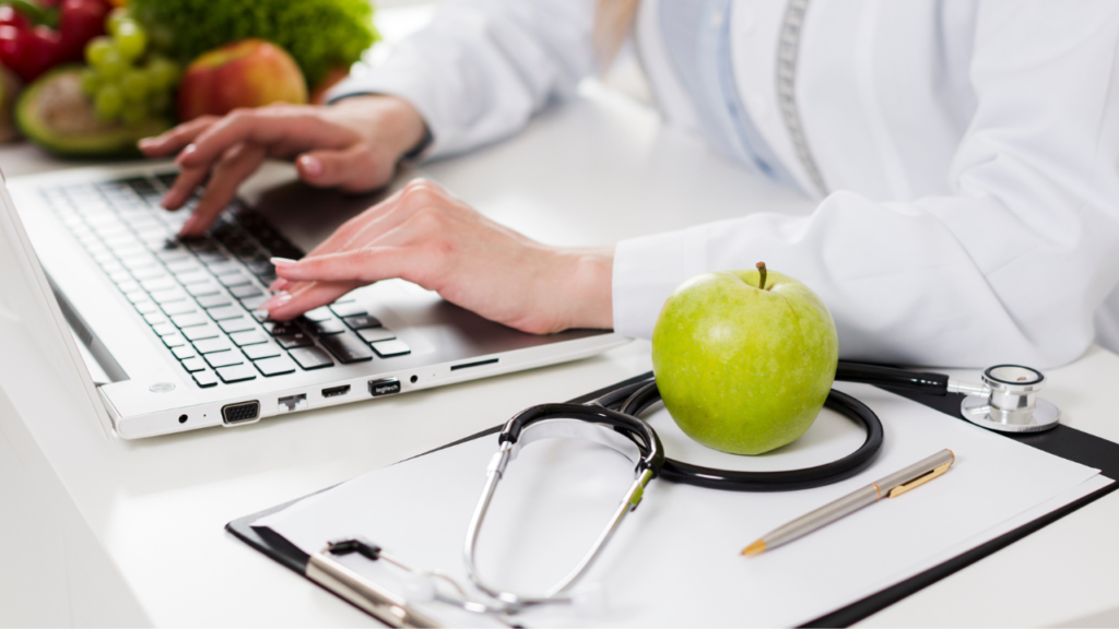 Uma nutricionista sentada na mesa do seu consultório, ao lado de um estetoscópio e uma maça verde /o que é nutricionista