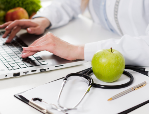 Uma nutricionista sentada na mesa do seu consultório, ao lado de um estetoscópio e uma maça verde /o que é nutricionista