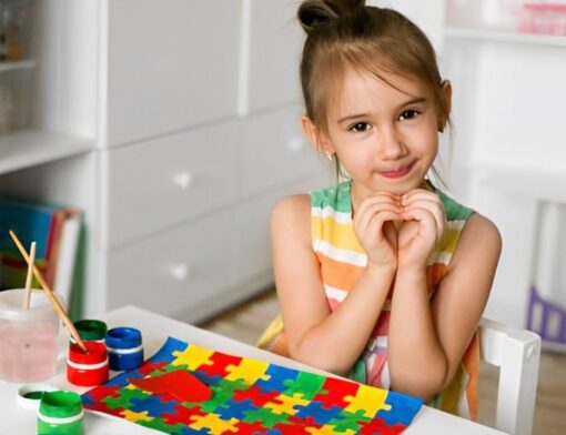 Menina autista com pintura de quebra-cabeça na mesa a sua frente enquanto ela faz coração com as mãos